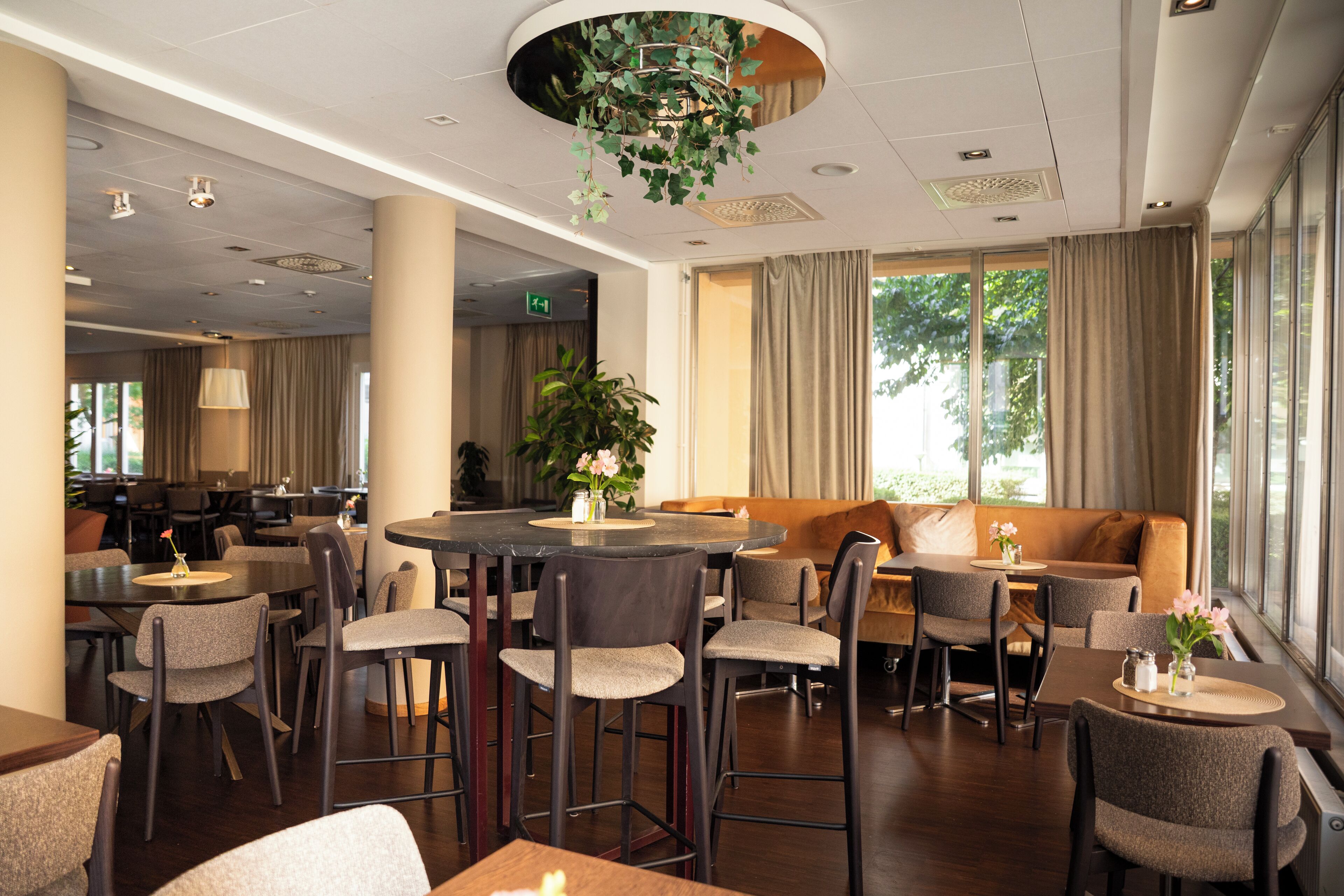 A hotel dining area with round tables, bar stools, and a large window with curtains.