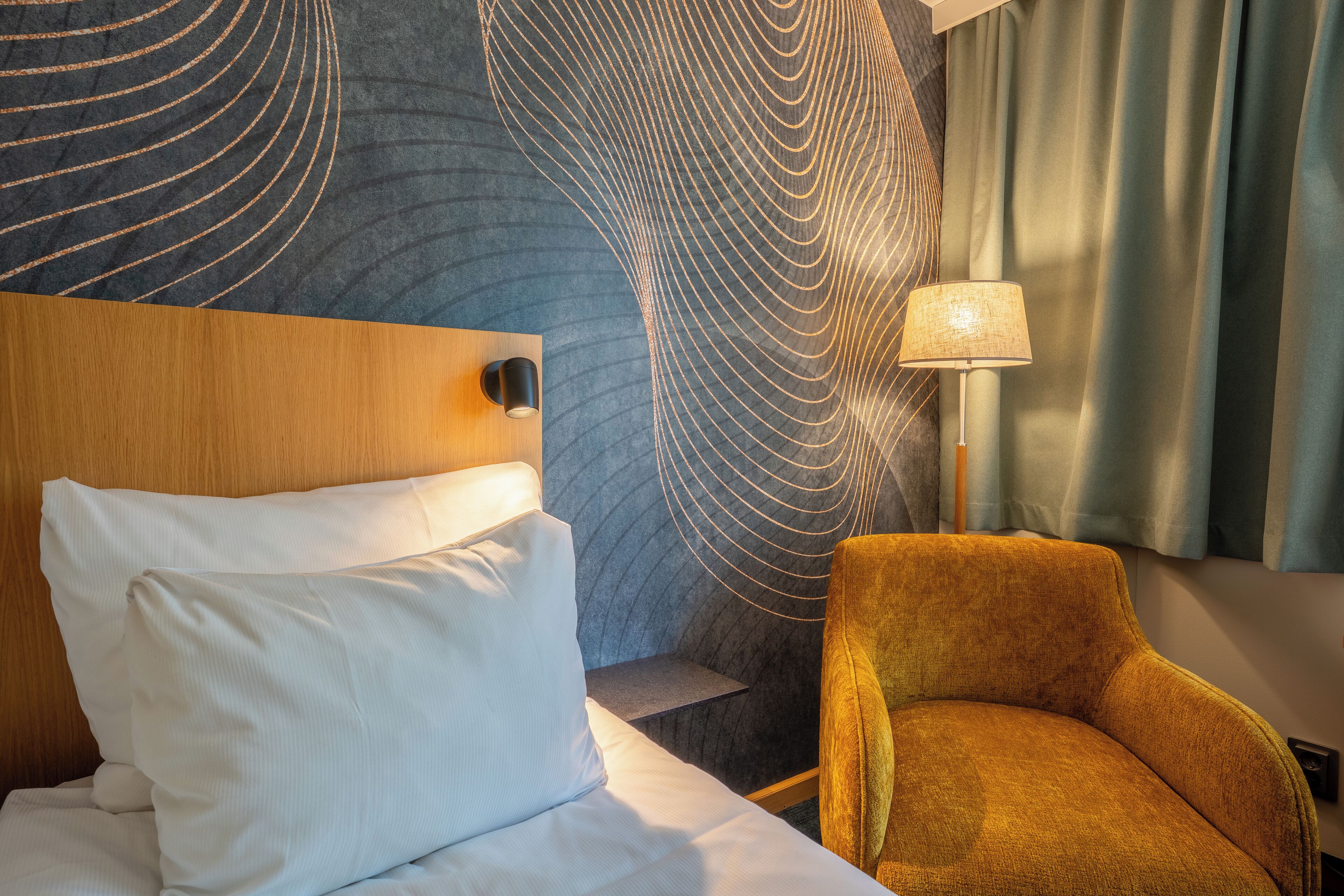 Una habitación de hotel con una cama, cabecera, pared con diseño geométrico, lámpara y una silla.