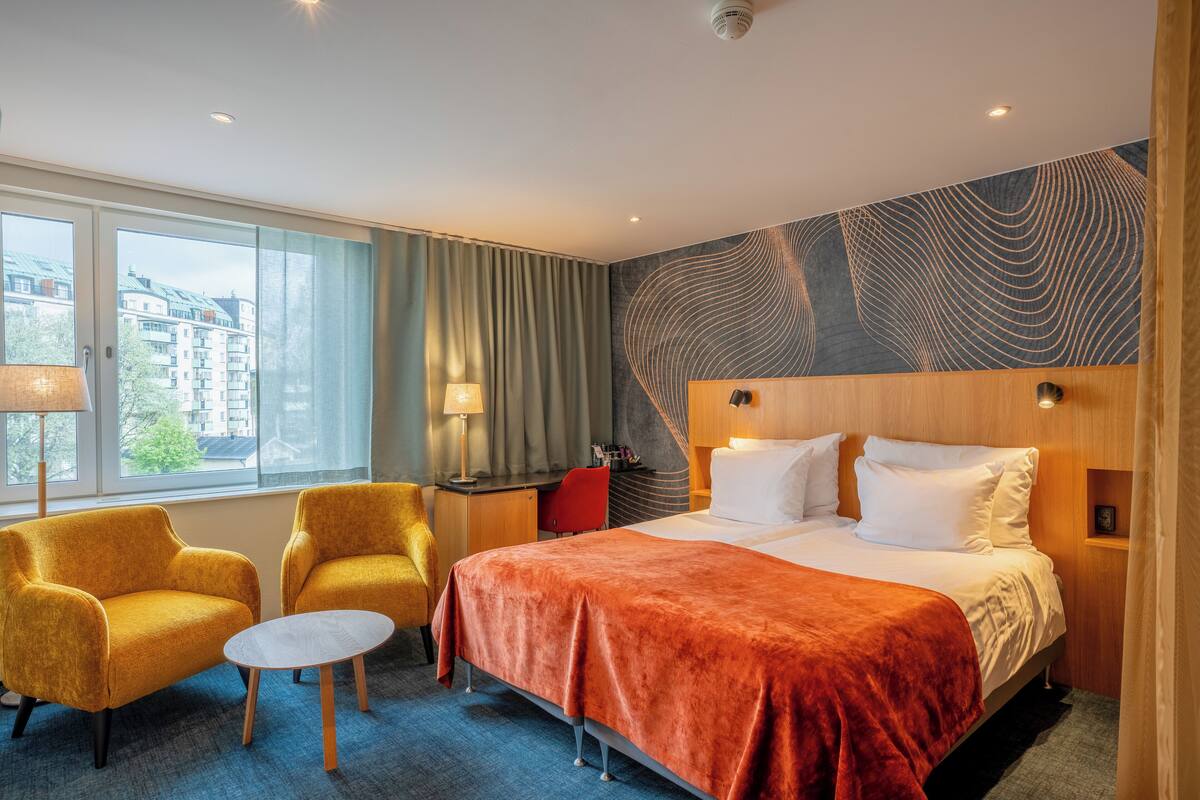 A hotel room with a bed, two yellow armchairs, a small table, and a view of buildings through large windows.