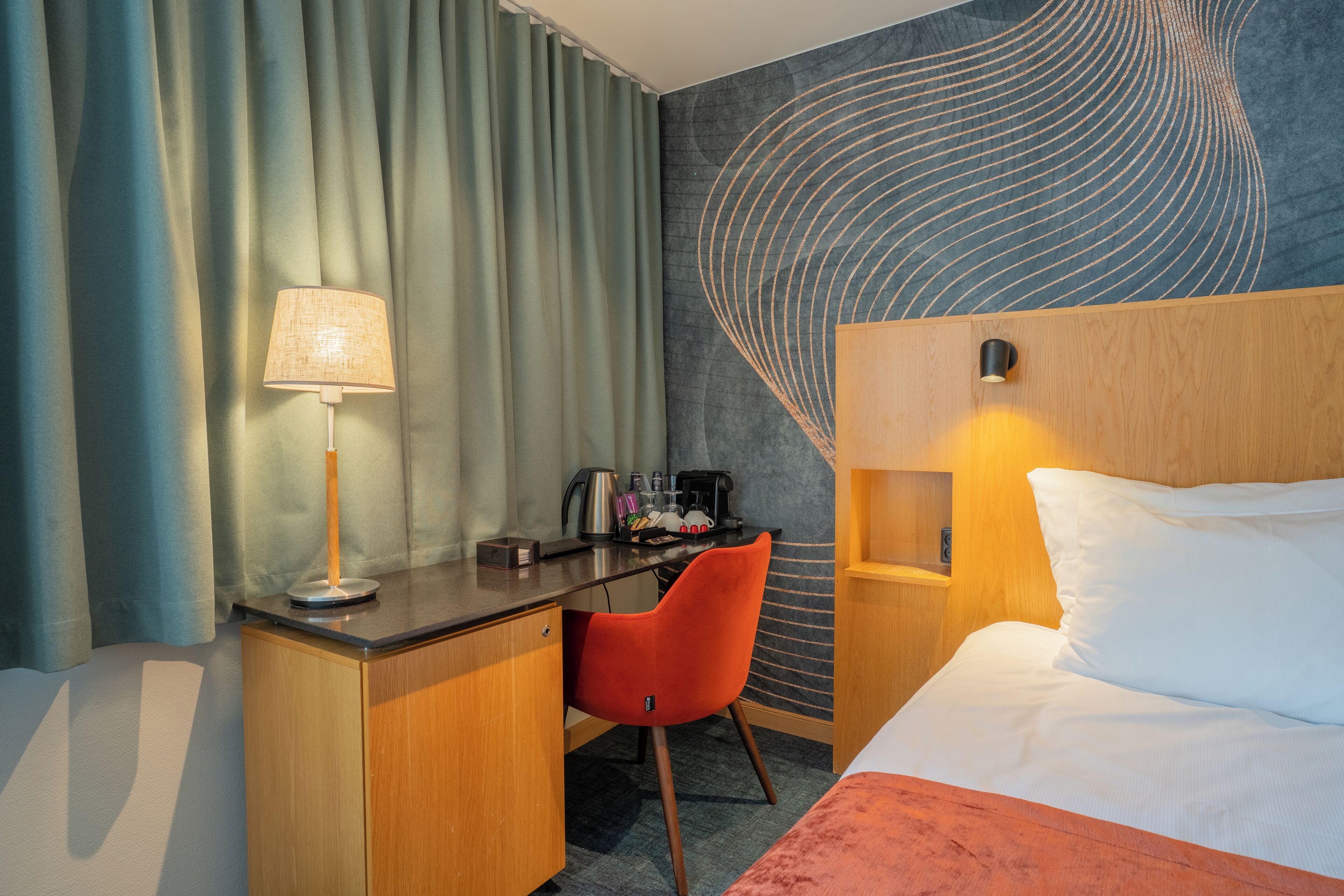 Habitación de hotel con cama, escritorio, silla, lámpara y una pared con diseño ondulado.