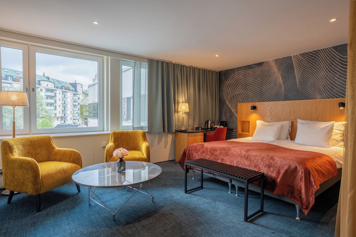 A hotel room with a bed, two armchairs, a round table with a vase, a bench, a desk with a lamp, and a view of buildings through large windows.