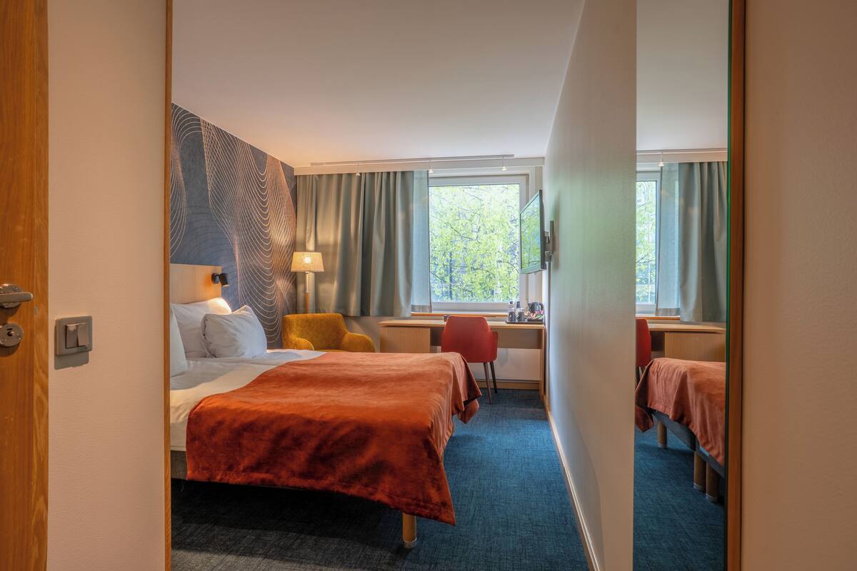 A hotel room with two beds, a desk, a chair, a lamp, and a large window.