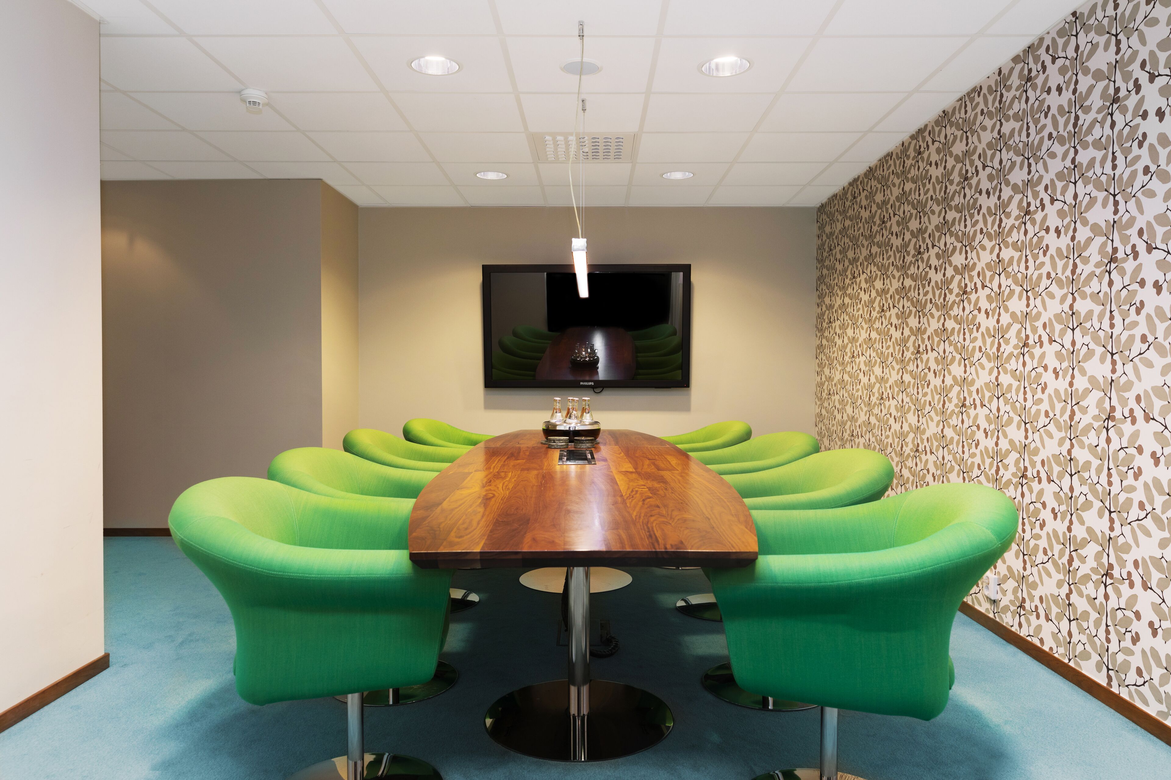 A conference room with a wooden table, green chairs, a flat-screen TV, and patterned wallpaper.