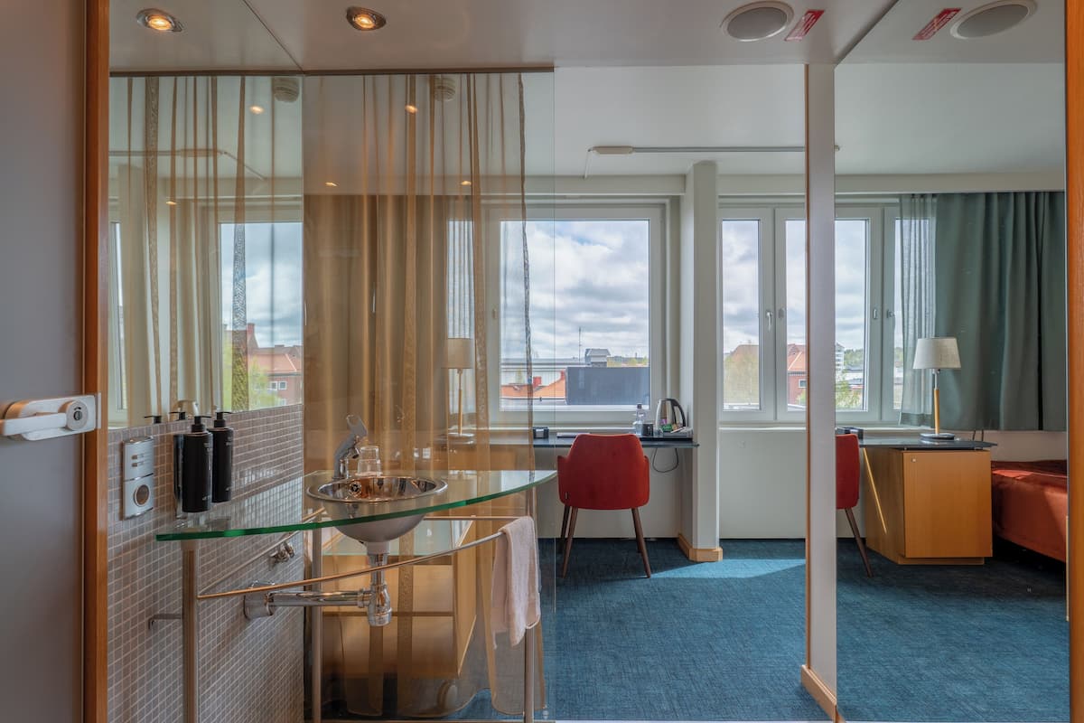 A hotel room with a glass shower enclosure, a desk with a chair, a bed, and a view of the cityscape through large windows.