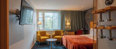Une chambre d’hôtel avec un lit, deux fauteuils jaunes, une petite table ronde et un téléviseur fixé au mur.
