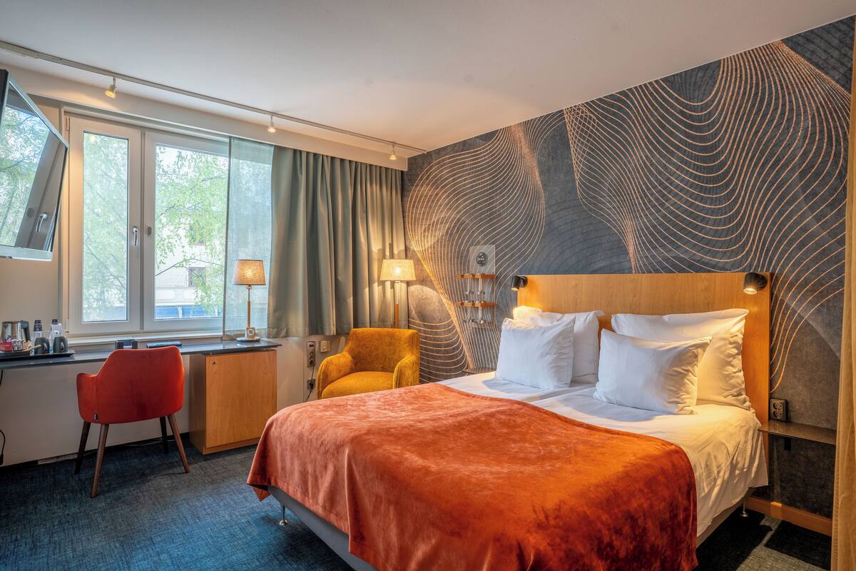 A hotel room with a large bed, a desk with a chair, and a view of trees through the window.