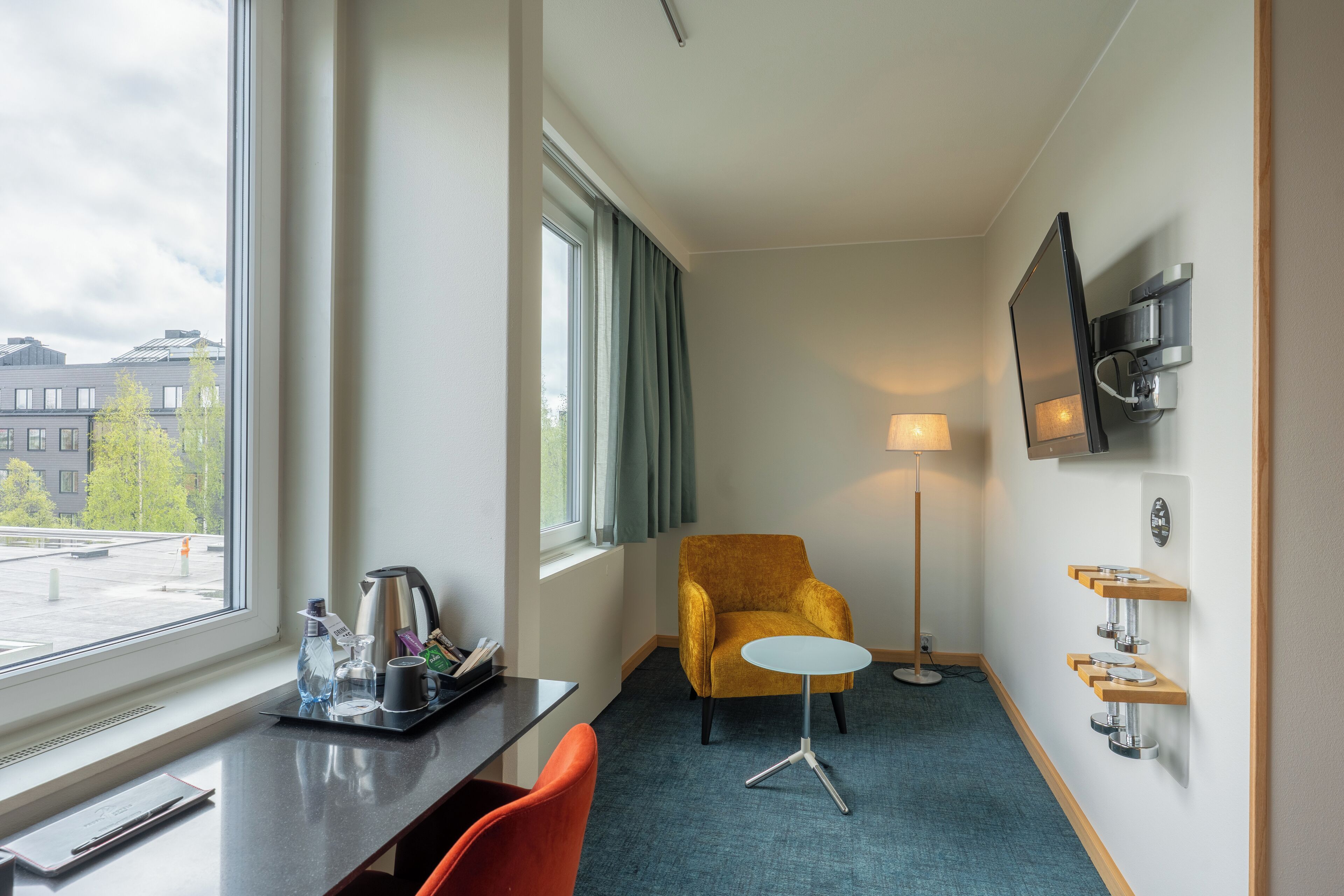 A hotel room with a desk, a chair, a sofa, a TV mounted on the wall, a floor lamp, and a window with curtains.