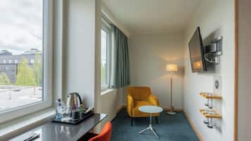 Een hotelkamer met een bureau, een stoel, een bank, een aan de muur bevestigde televisie, een vloerlamp en een raam met gordijnen.