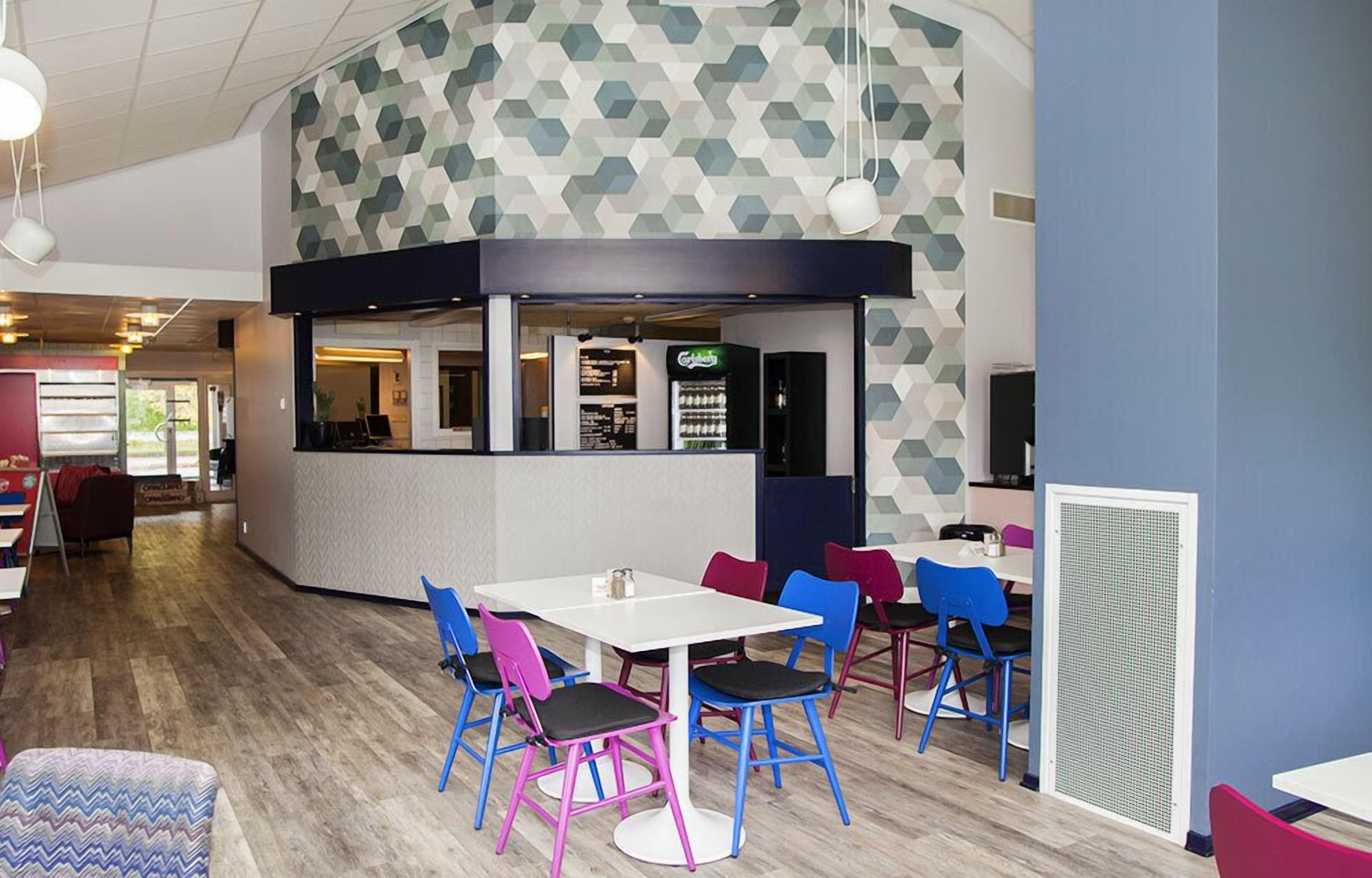 A modern indoor space with a central white table surrounded by colorful chairs, a counter with a beverage dispenser, and a geometric patterned wall.