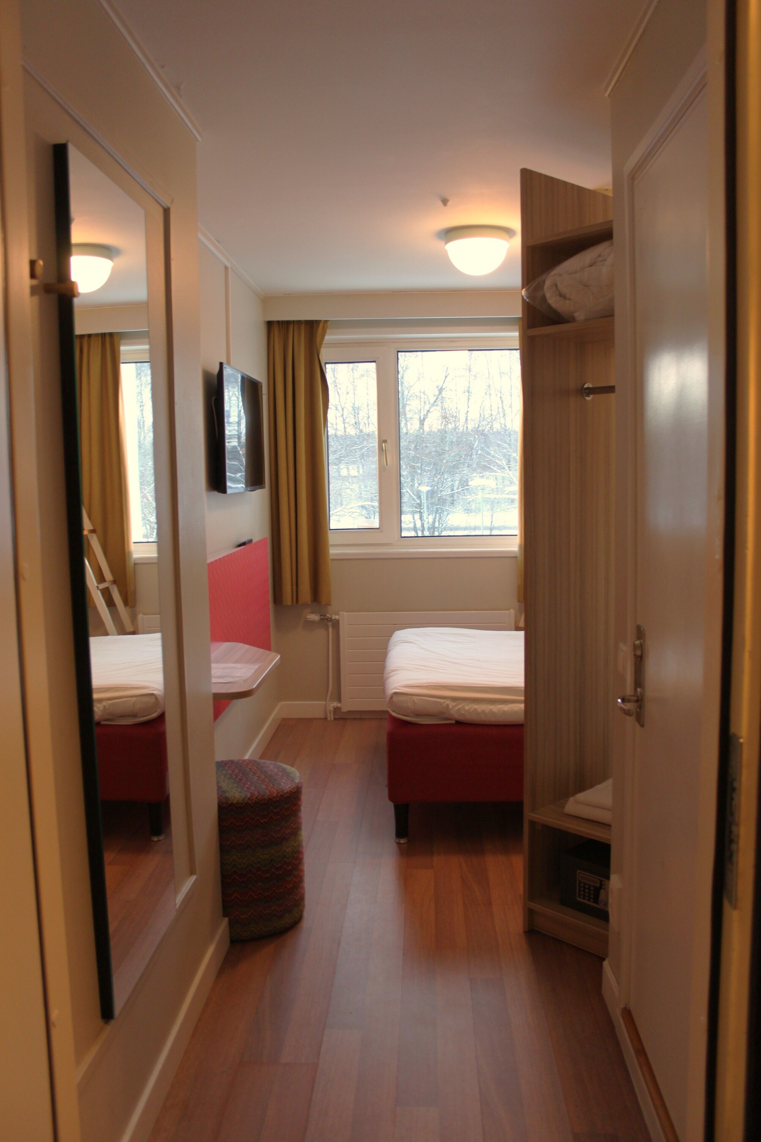 A narrow hotel room with two beds, a wardrobe, a mirror, and a window with curtains.
