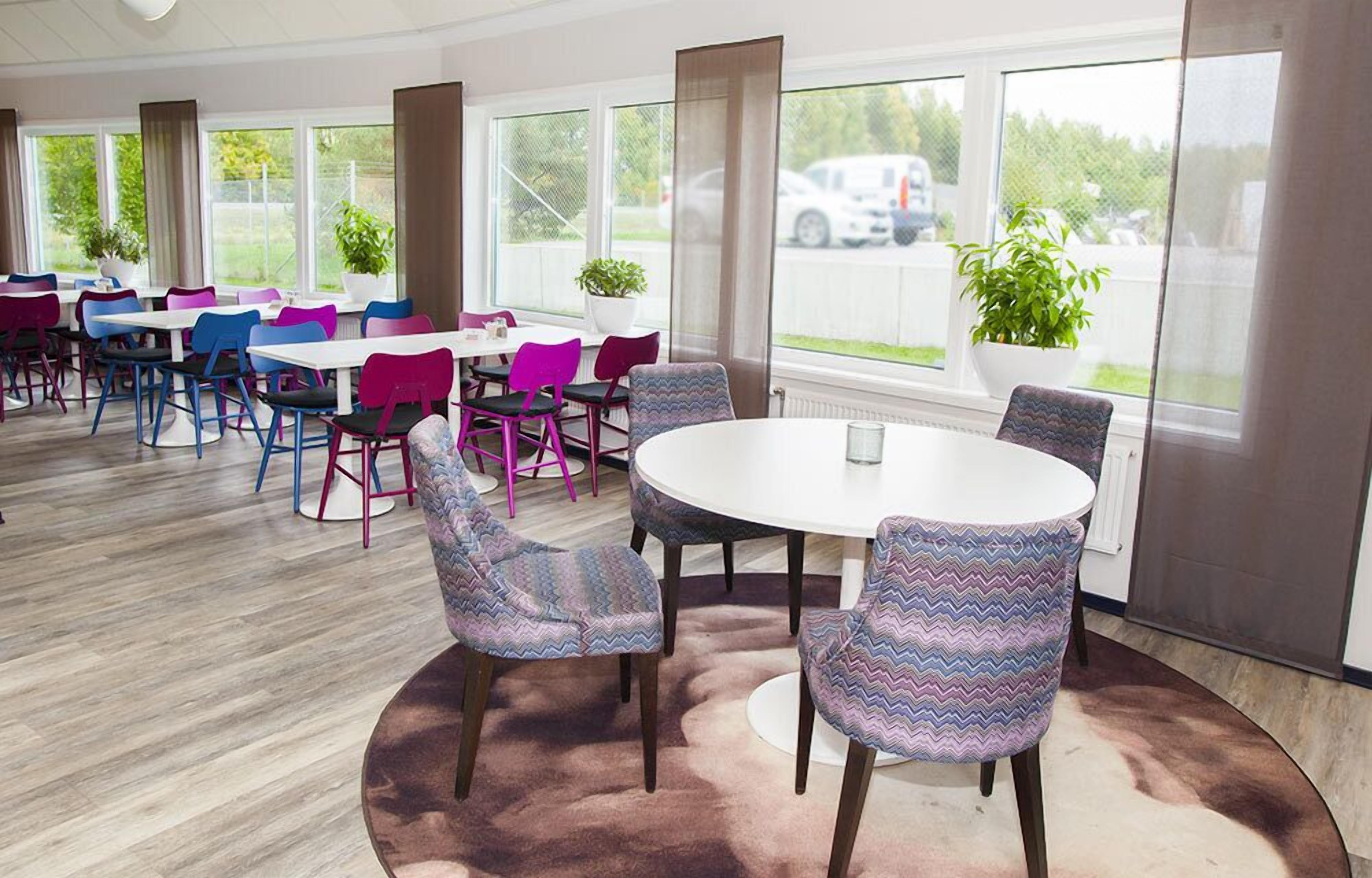 A dining area with round tables and colorful chairs, large windows with curtains, and potted plants.