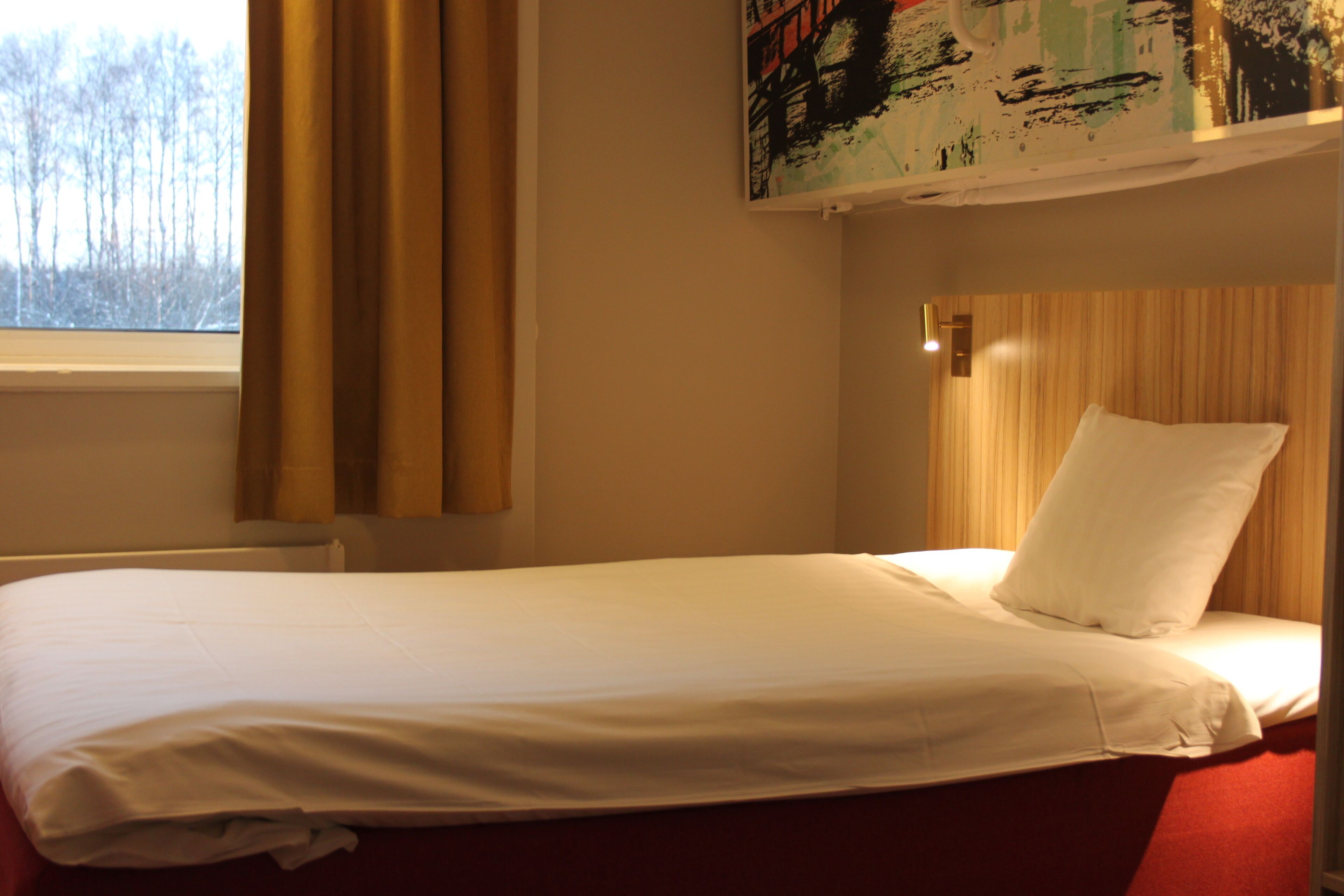 A single bed with white bedding in a room with a window, a wooden headboard, and a wall-mounted artwork.