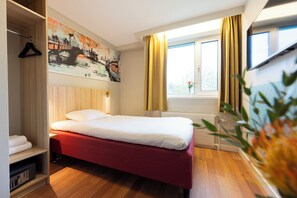 Une chambre d’hôtel avec un lit, un tableau au mur, une fenêtre avec des rideaux et une plante.