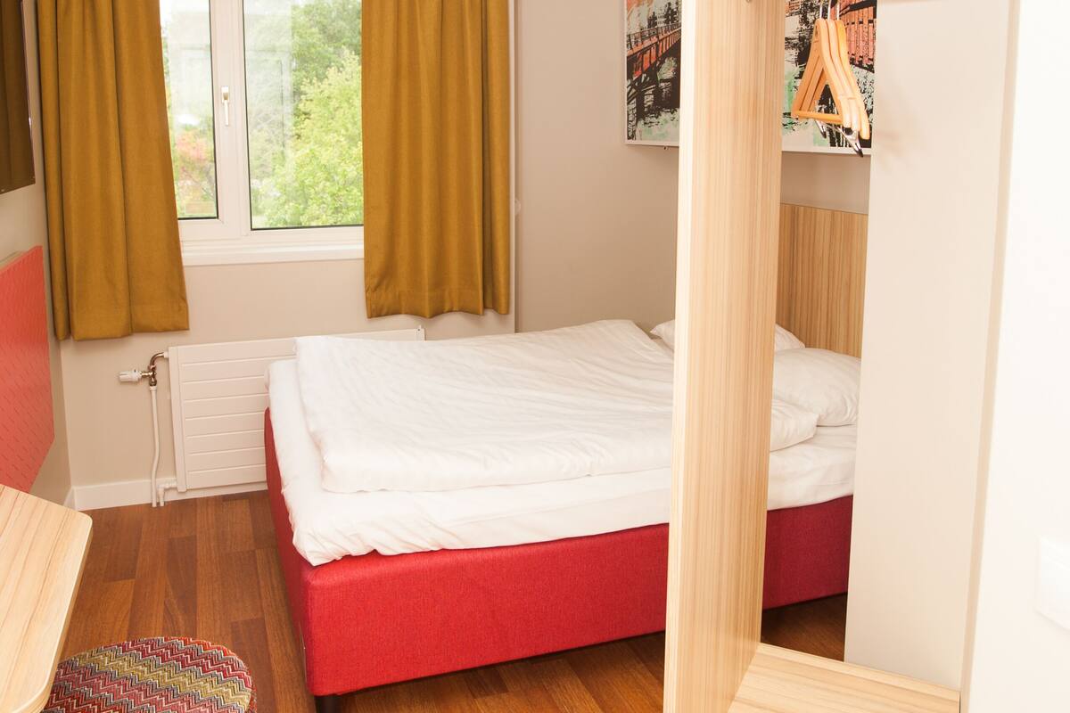 A bedroom with a bed, a wardrobe, a radiator, a window with curtains, and a small ottoman.