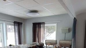 A corner room with a kitchenette, a window with curtains, a ceiling light, and a lamp on the wall.