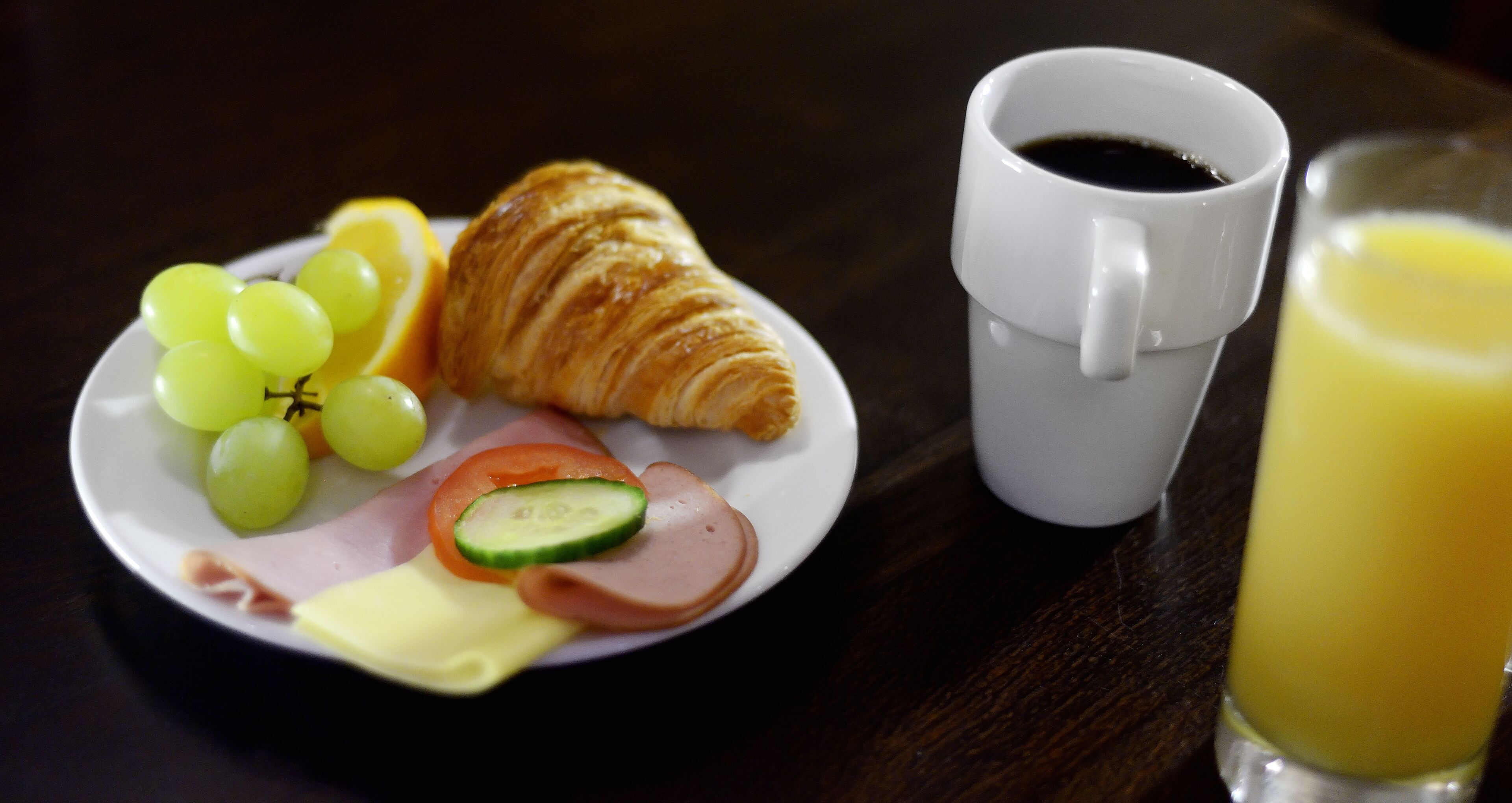 Un plato con jamón, queso, tomates, pepinos y uvas, un croissant, una taza de café y un vaso de jugo de naranja sobre una mesa.