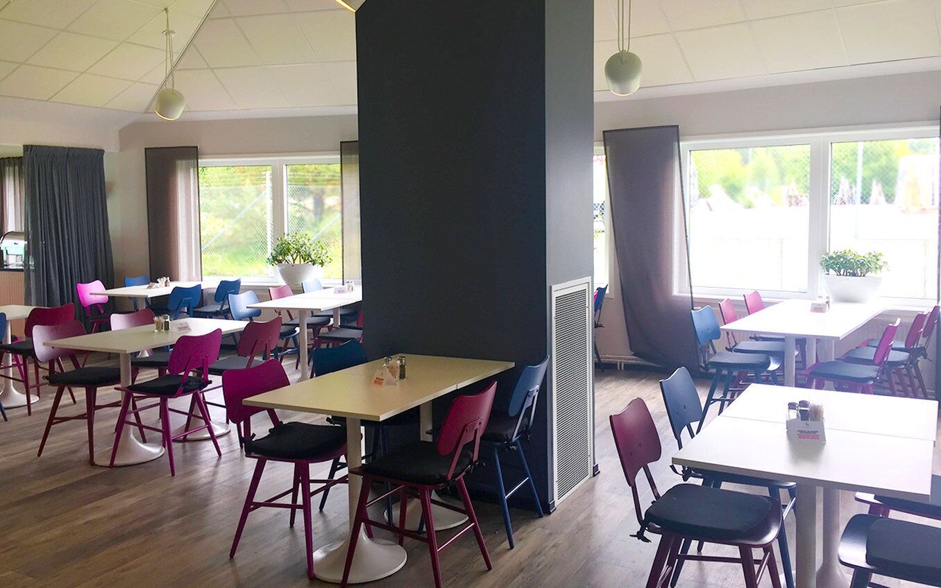 A dining area with multiple tables and chairs, large windows with views of greenery, and a central pillar.