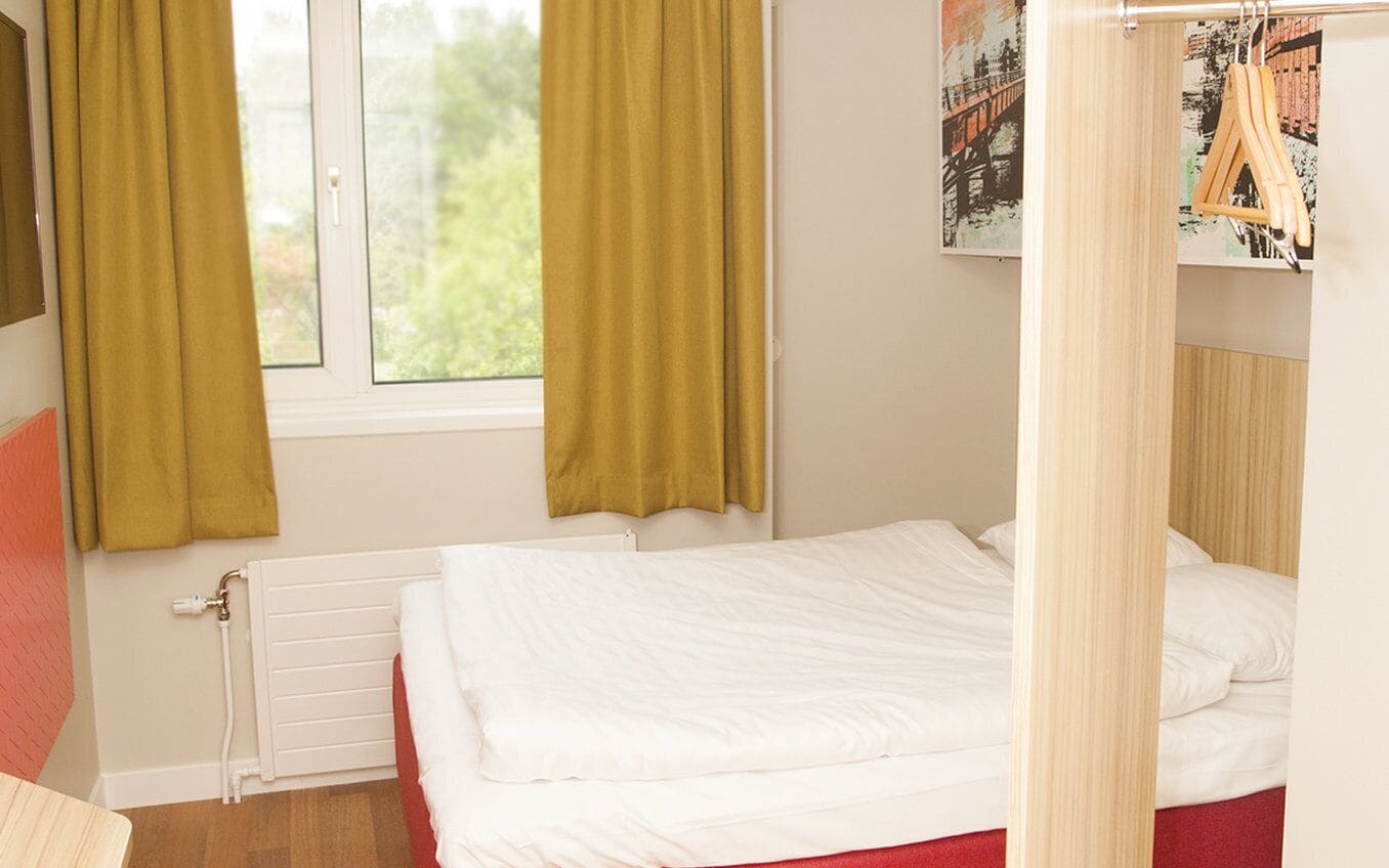 A bedroom with a bed, a radiator, a window with curtains, and a wall-mounted artwork.