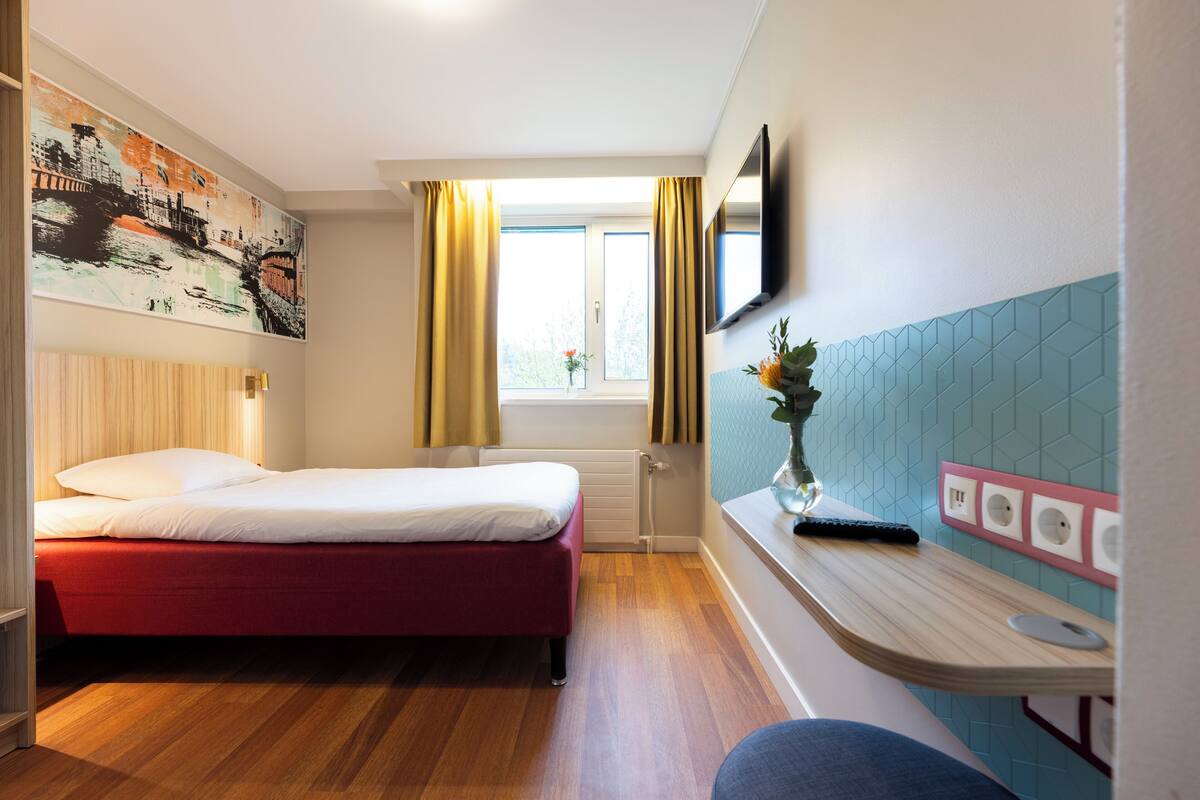 A hotel room with a bed, a desk, a window with curtains, a painting on the wall, and a tiled wall.