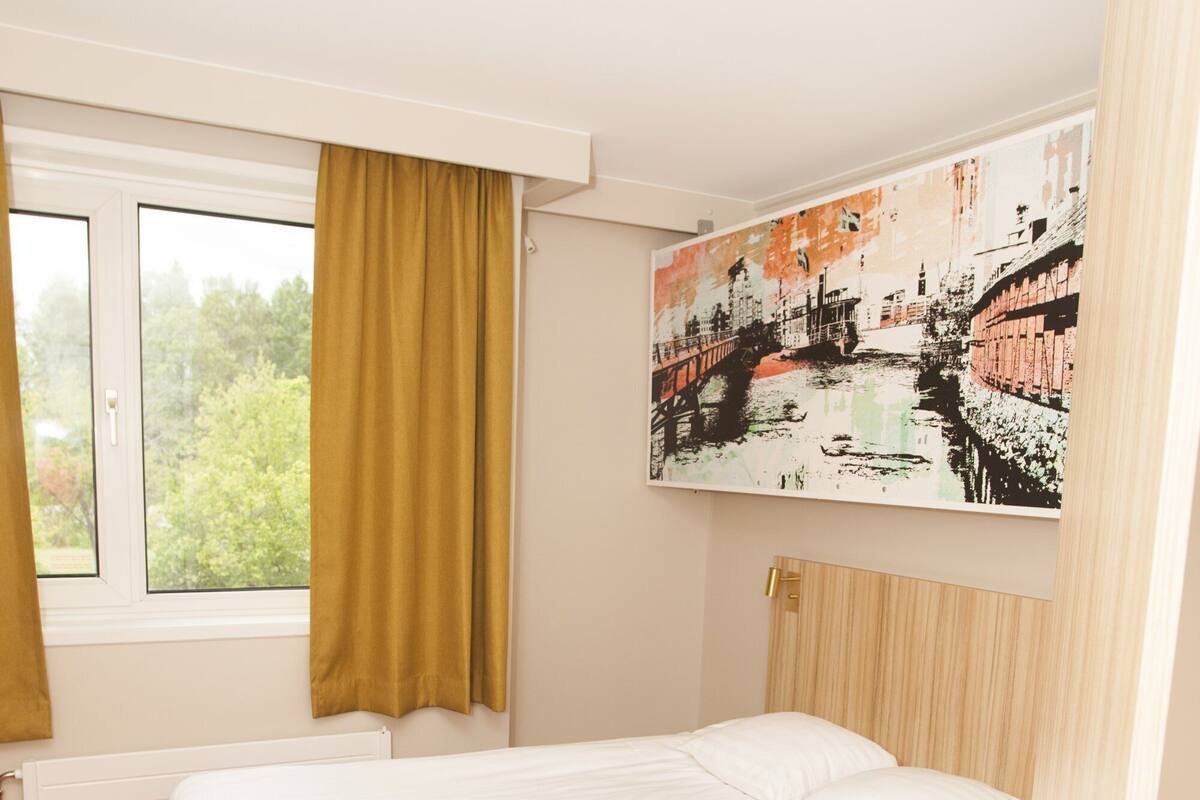 A bedroom with a red bed, a window with curtains, and a cityscape painting on the wall.