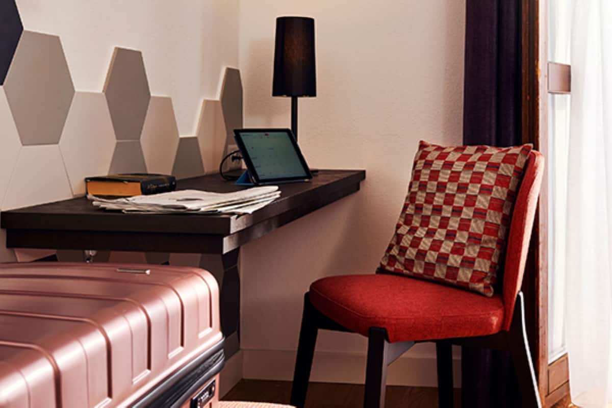 Une chambre d’hôtel avec un bureau, un ordinateur portable, une chaise rouge avec un coussin à carreaux et une valise marron.