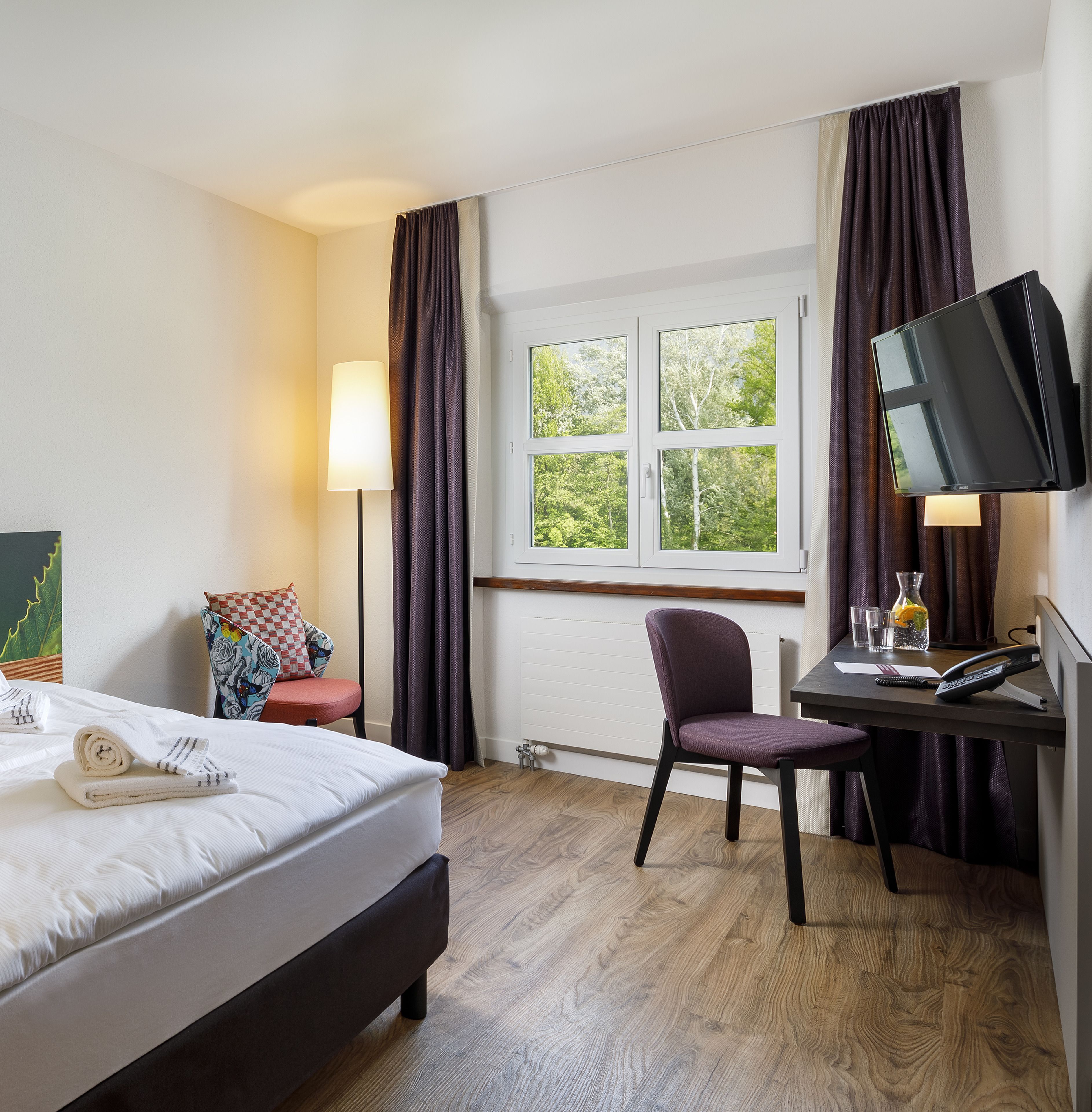Una habitación de hotel con una cama, un escritorio con computadora y teléfono, una silla, una ventana con cortinas y una lámpara.