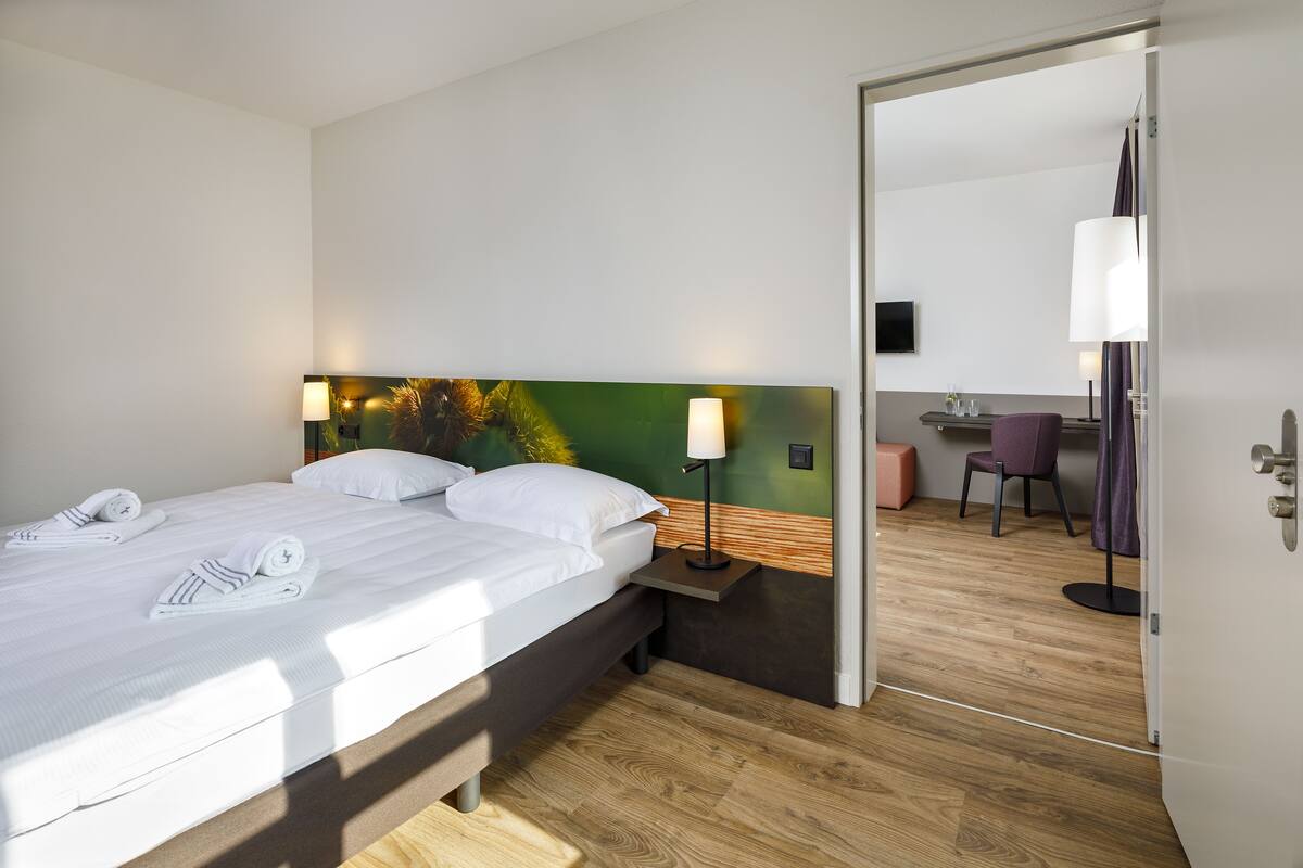 Une chambre d’hôtel avec un lit bien fait, une lampe de chevet, une tête de lit en bois et une vue sur la salle de bain par une porte ouverte.