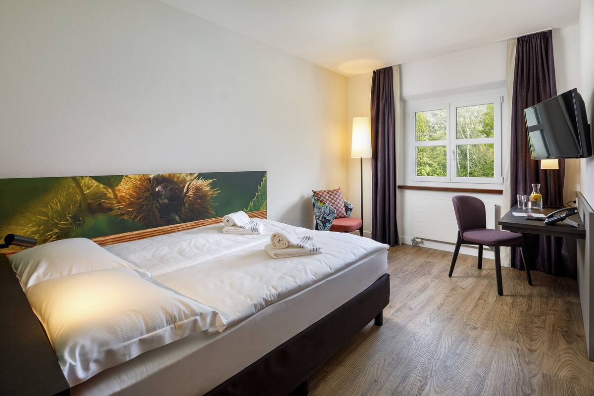 Une chambre d’hôtel comprenant un lit, un bureau avec une chaise, une télévision et une fenêtre avec des rideaux.