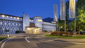 A modern hotel with a curved entrance, multiple flags, and a well-lit facade. - Hotel Bellinzona Sud Swiss Quality (Bellinzona)