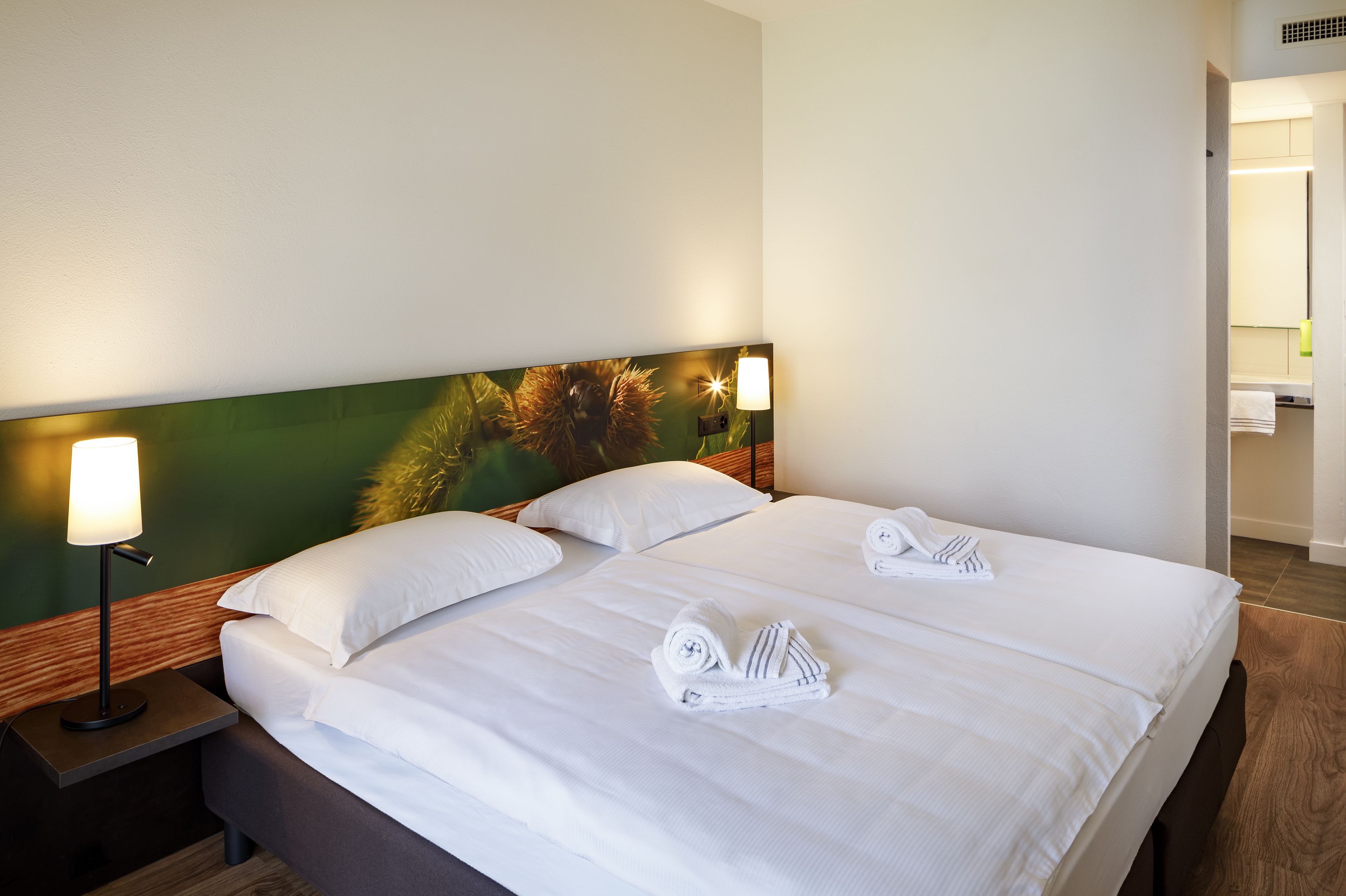 Quarto de hotel com uma cama grande, duas almofadas brancas, mesa de cabeceira com abajur e cabeceira com padrão verde e marrom.