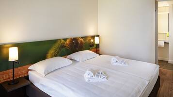 Quarto de hotel com uma cama grande, duas almofadas brancas, mesa de cabeceira com abajur e cabeceira com padrão verde e marrom.