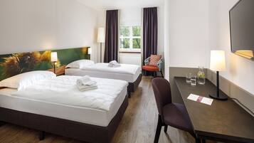 Quarto de hotel com duas camas, uma escrivaninha e uma cadeira. Há uma janela com cortinas, um quadro na parede e uma luminária na escrivaninha.