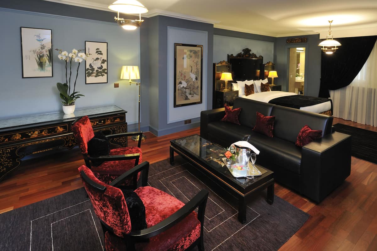 A modern hotel room with a black sofa, red velvet chairs, a glass coffee table, and artwork on the walls.