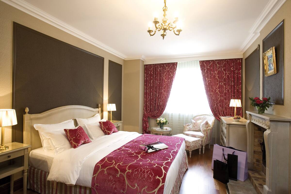 A well-appointed hotel room with a large bed, a chandelier, a fireplace, and a seating area with a chair and a small table.
