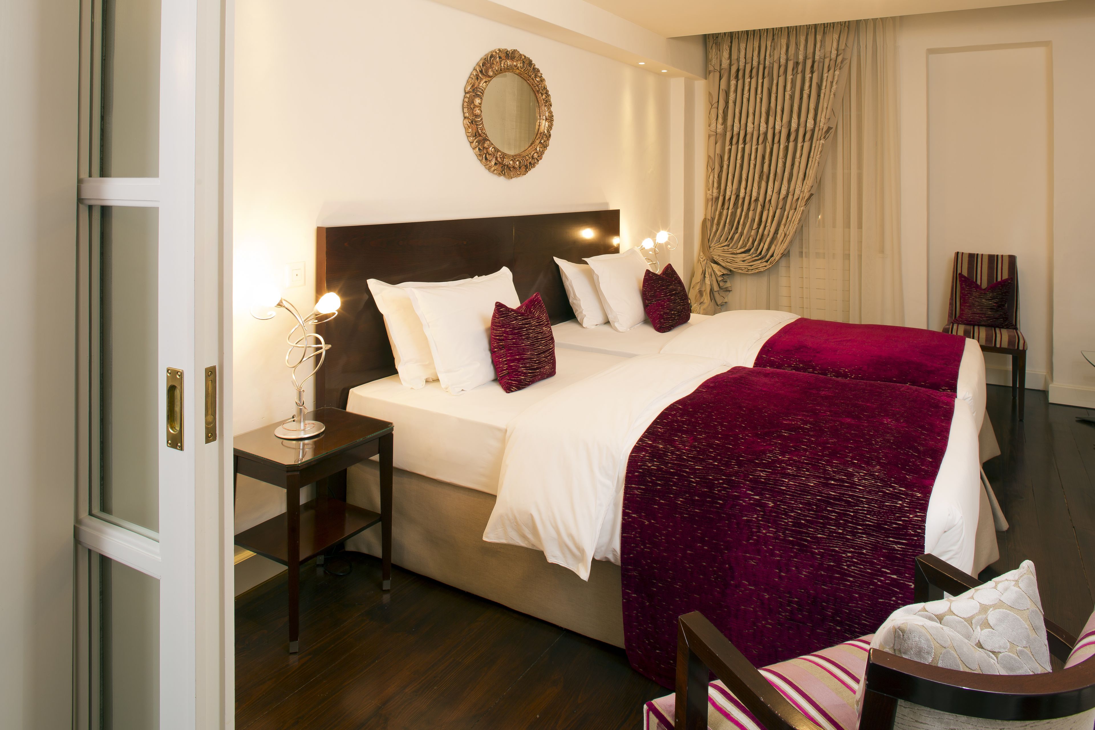 Quarto de hotel com uma cama grande, uma mesa de cabeceira com abajur, uma cadeira e um espelho na parede.