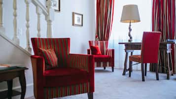 Una sala de estar con un sillón rojo, un sillón a rayas y una butaca roja. Hay una lámpara sobre una mesa, y se ve una escalera al fondo.