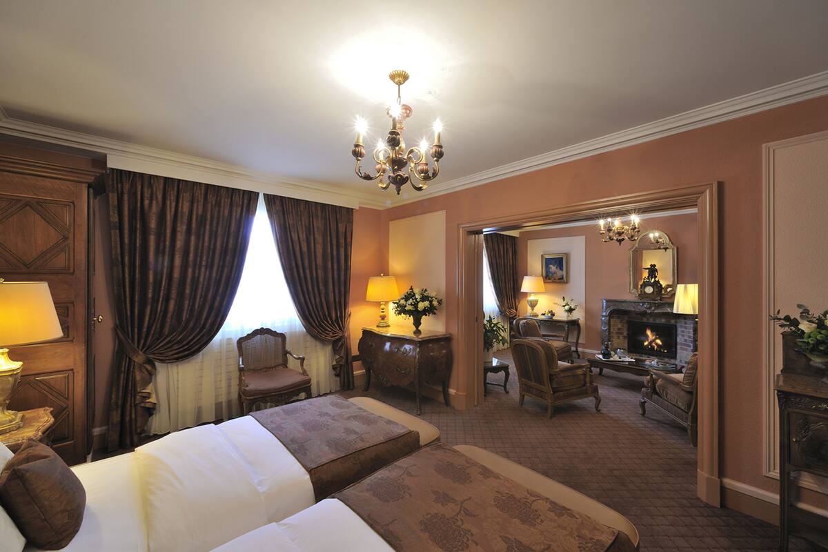 A hotel room with a large bed, a desk, a chair, a fireplace, and a chandelier.