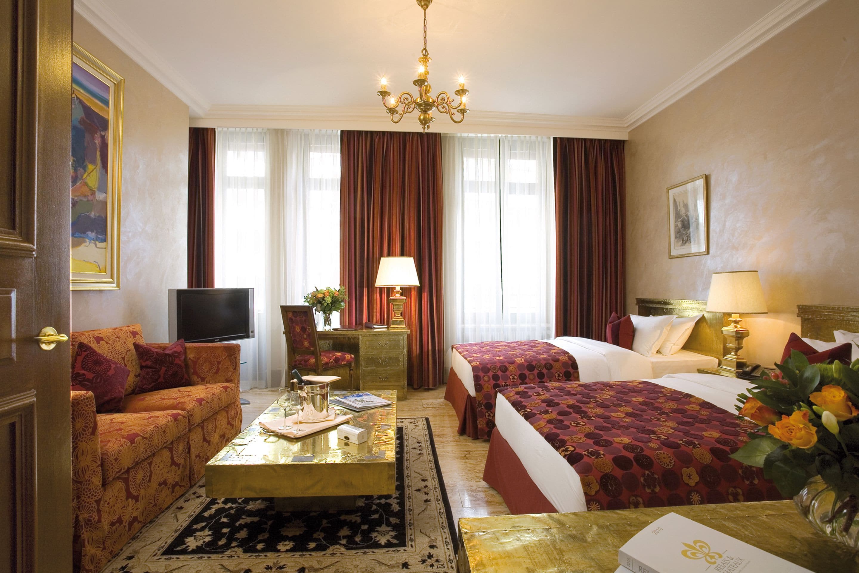 Una habitación de hotel con sofá, dos camas, televisor, escritorio con lámpara y candelabro.
