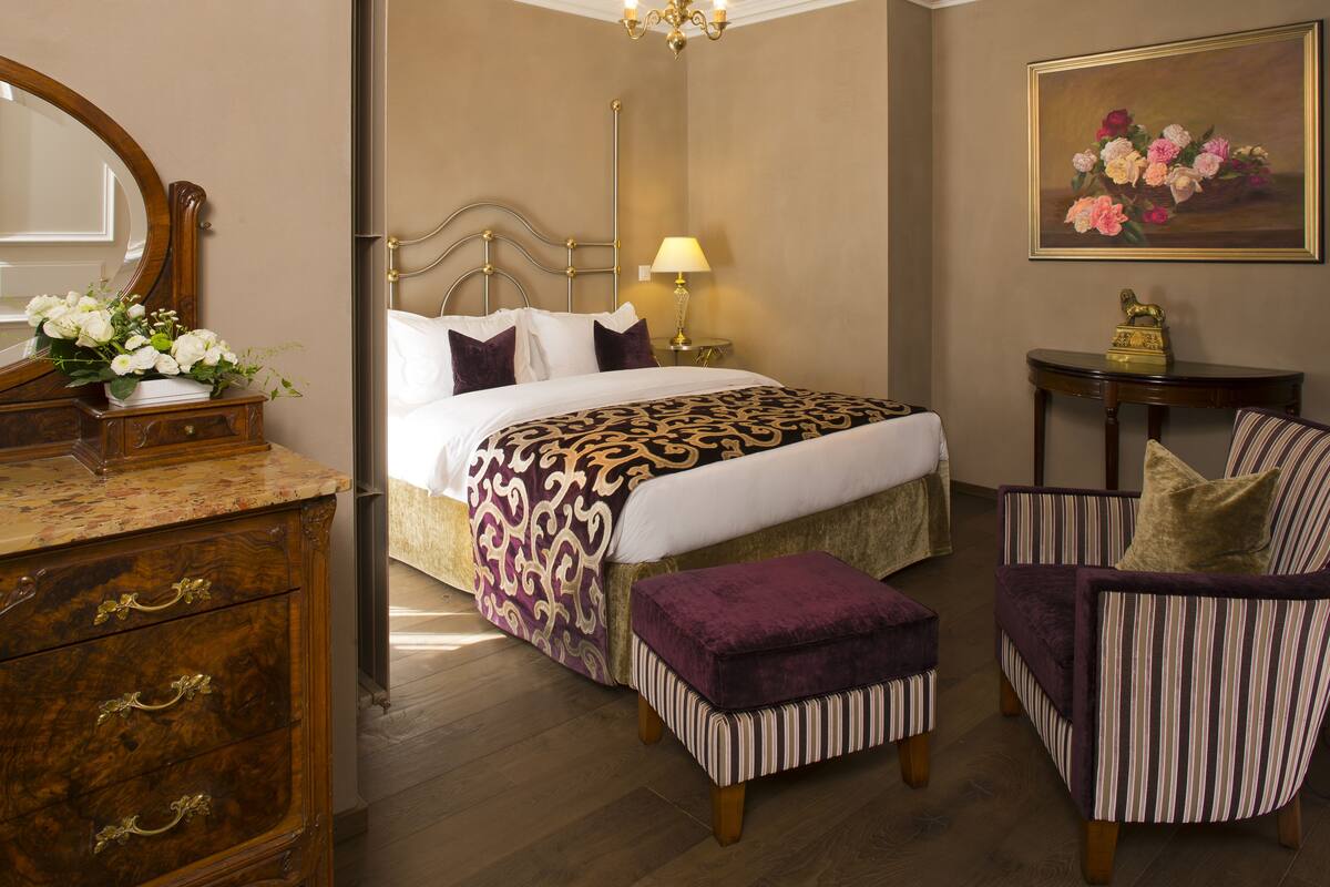 A hotel room with a bed, a dresser, a chair, and a small table with a lamp.