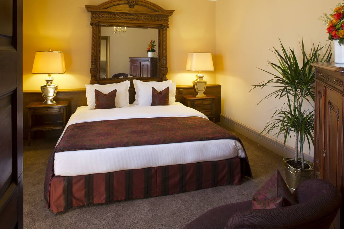A neatly made bed with a red and white bedspread, two bedside lamps, a large mirror above the bed, and a potted plant on the right.