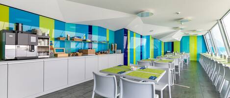 A modern dining area with white tables and chairs, a kitchen counter with appliances, and a colorful wall.