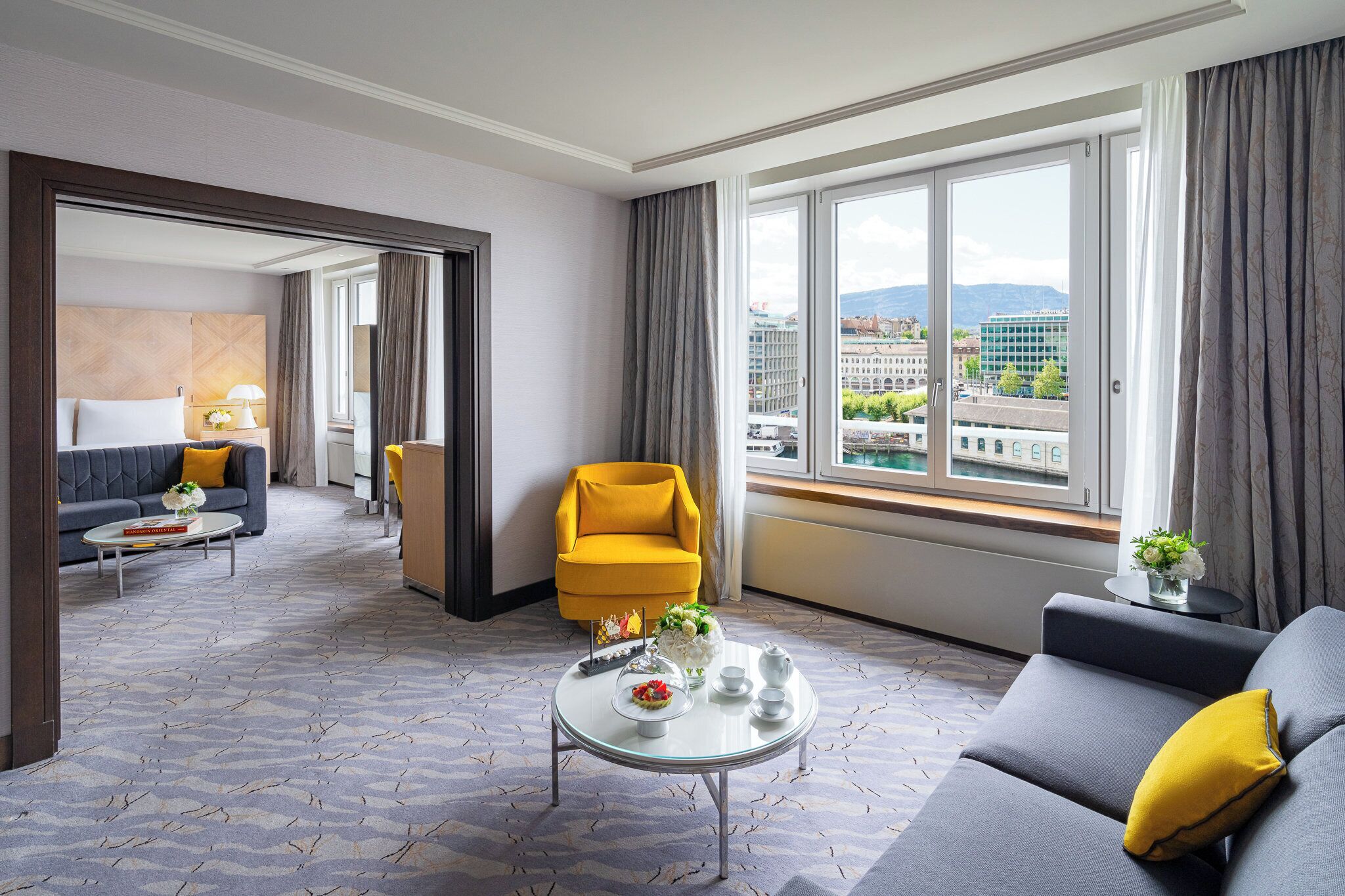 A modern hotel room with a sofa, armchair, and a round coffee table with a floral arrangement.