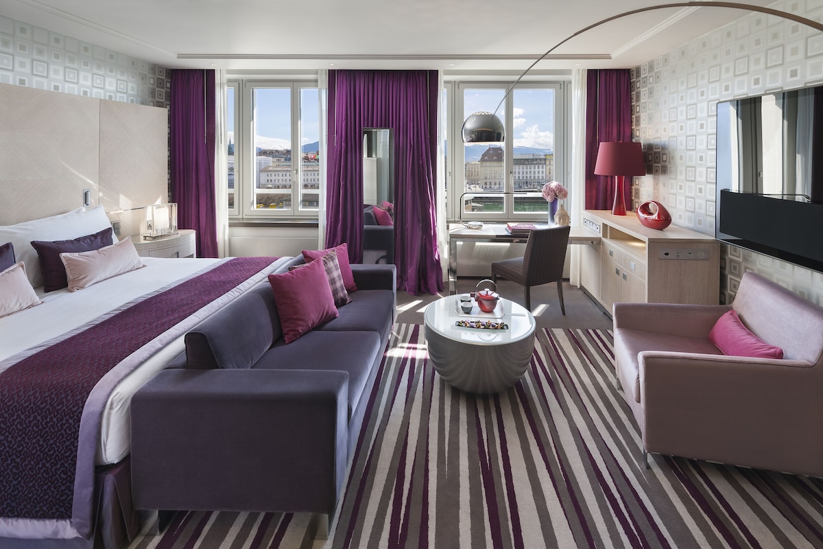A hotel room with a bed, a sofa, a desk, a chair, a TV, and a view of the city.