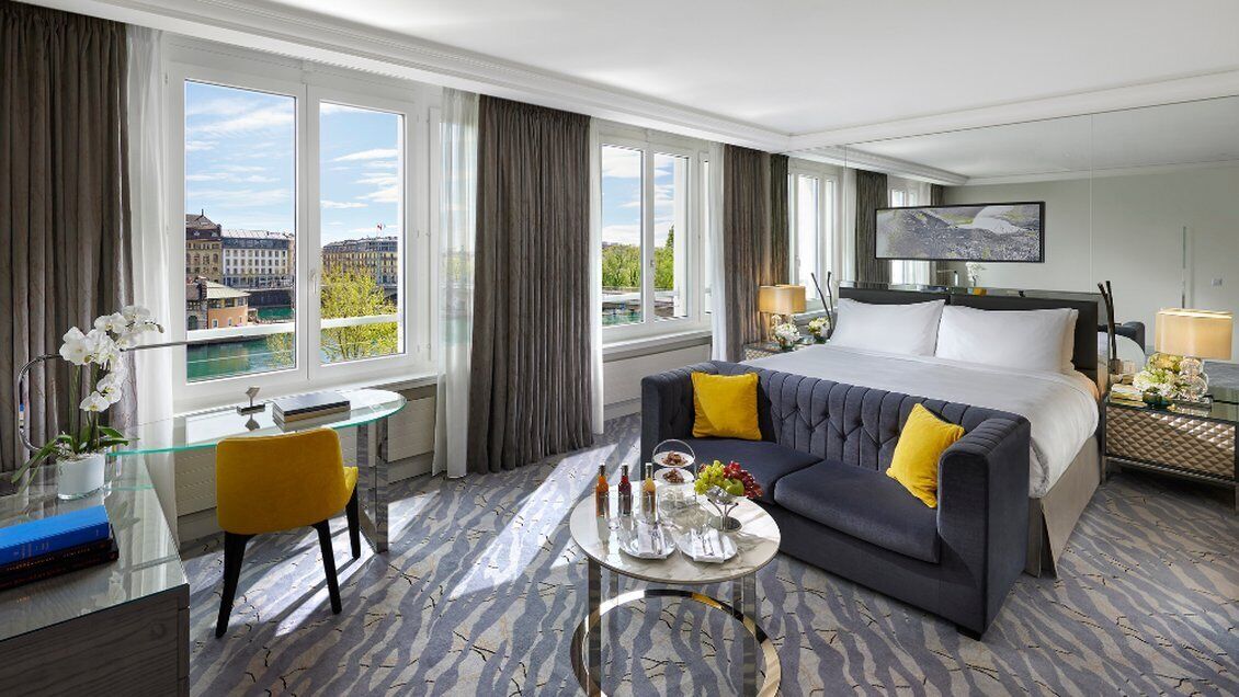 A hotel room with a bed, a sofa, a coffee table, a TV, and a view of buildings through large windows.