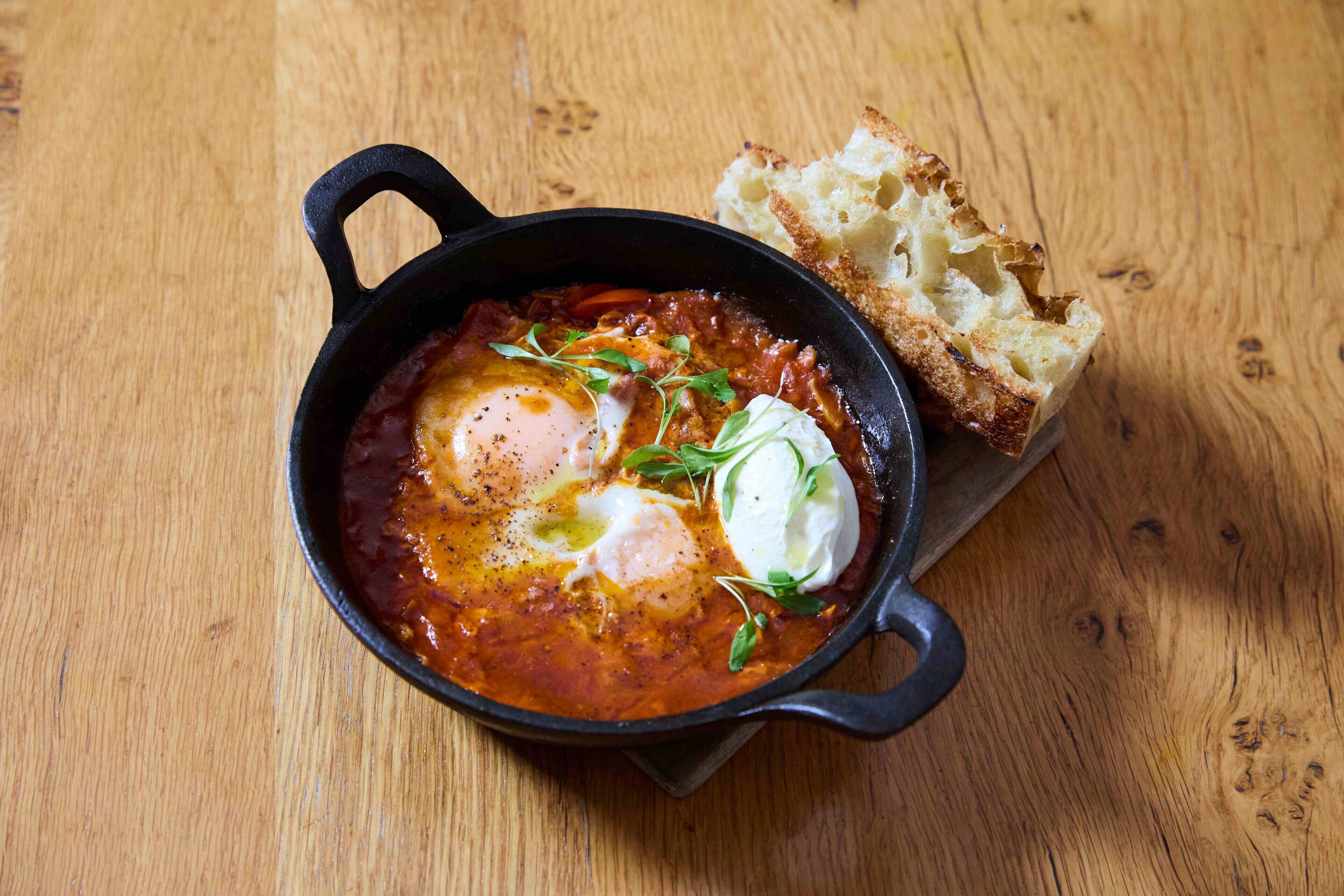 A cast iron skillet with a dish of eggs and sauce, accompanied by a piece of bread on a wooden table.