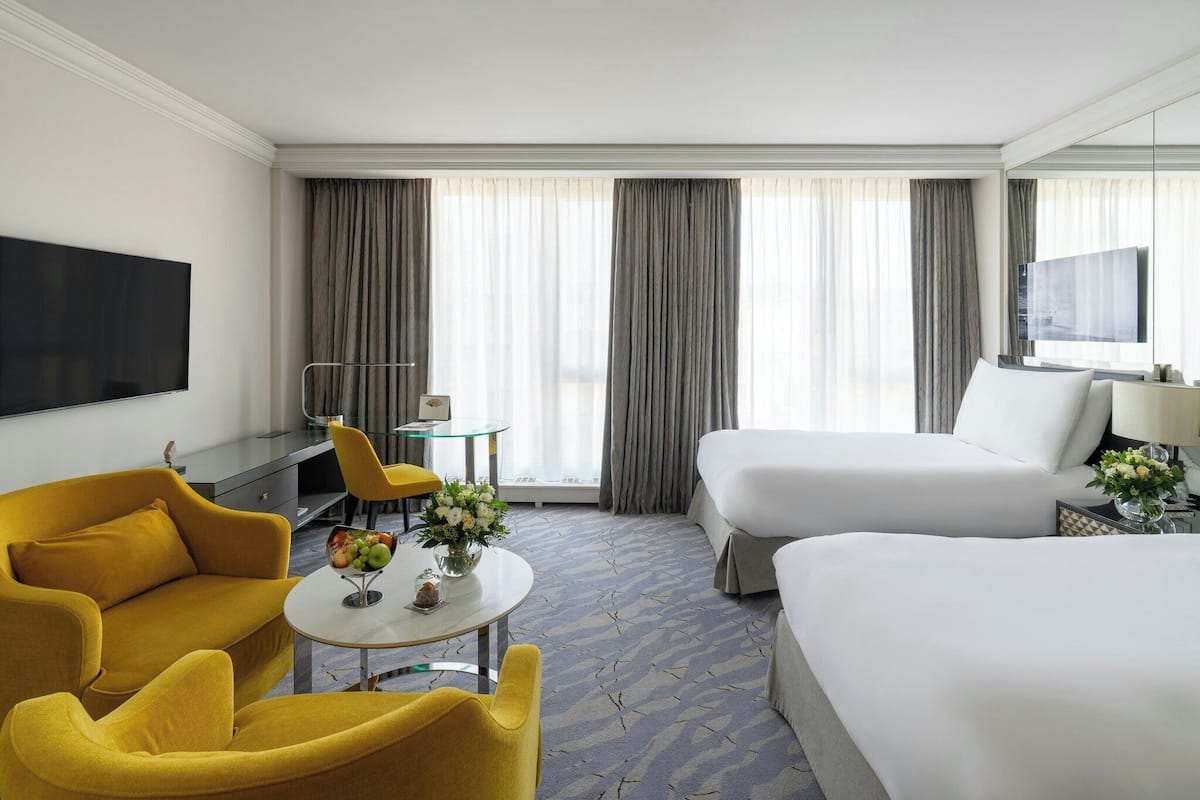 Habitación de hotel con cama, sofá, sillón, mesita con un florero, televisor y ventana con cortinas.