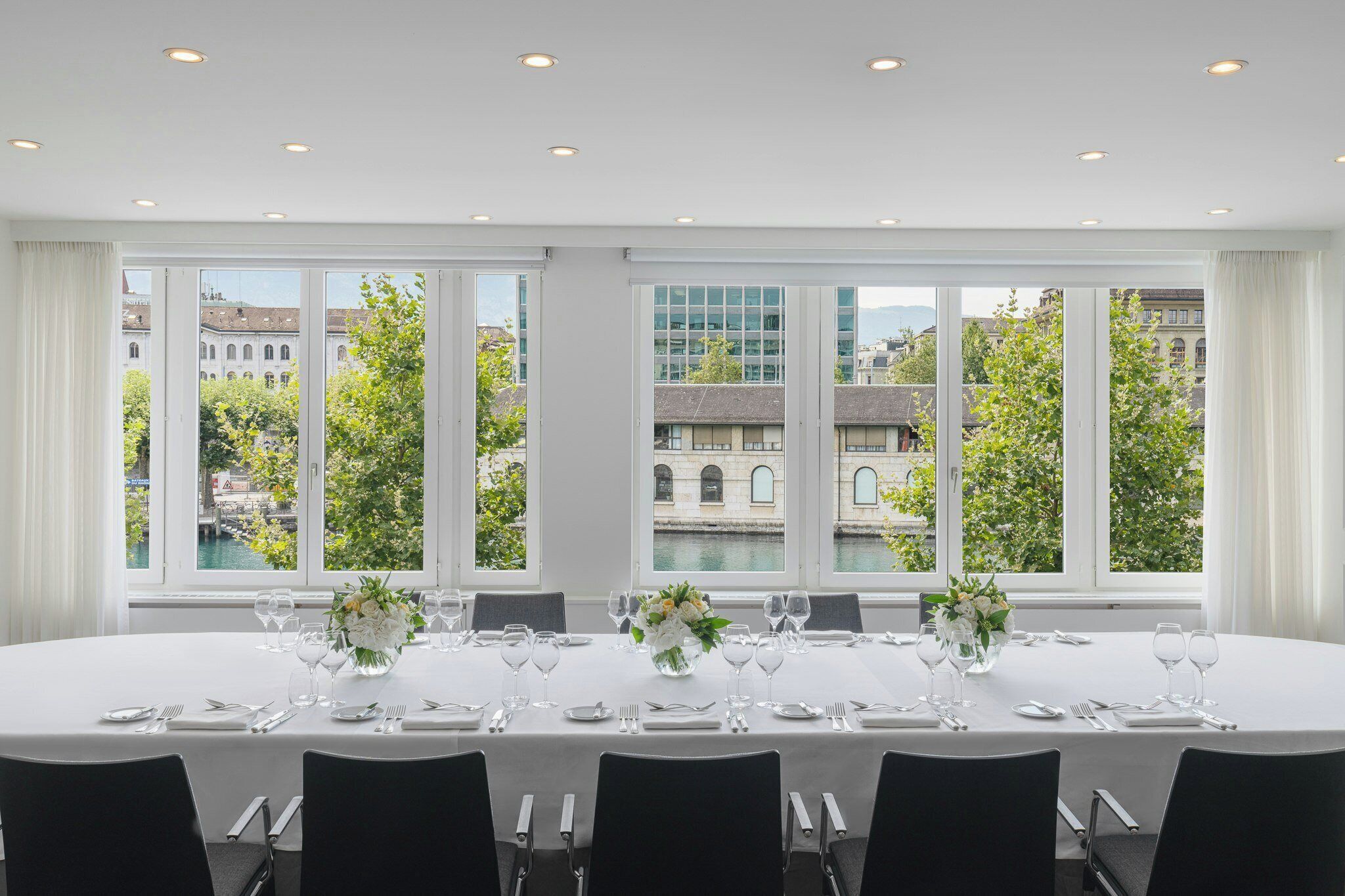 A dining room with a table set for six, large windows, and a view of buildings and trees.