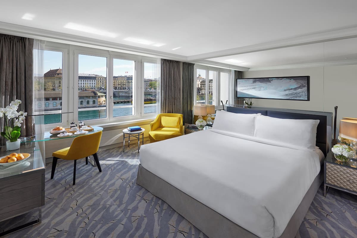 Habitación de hotel con una cama grande, un escritorio con silla, televisión y vistas a la ciudad a través de amplios ventanales.
