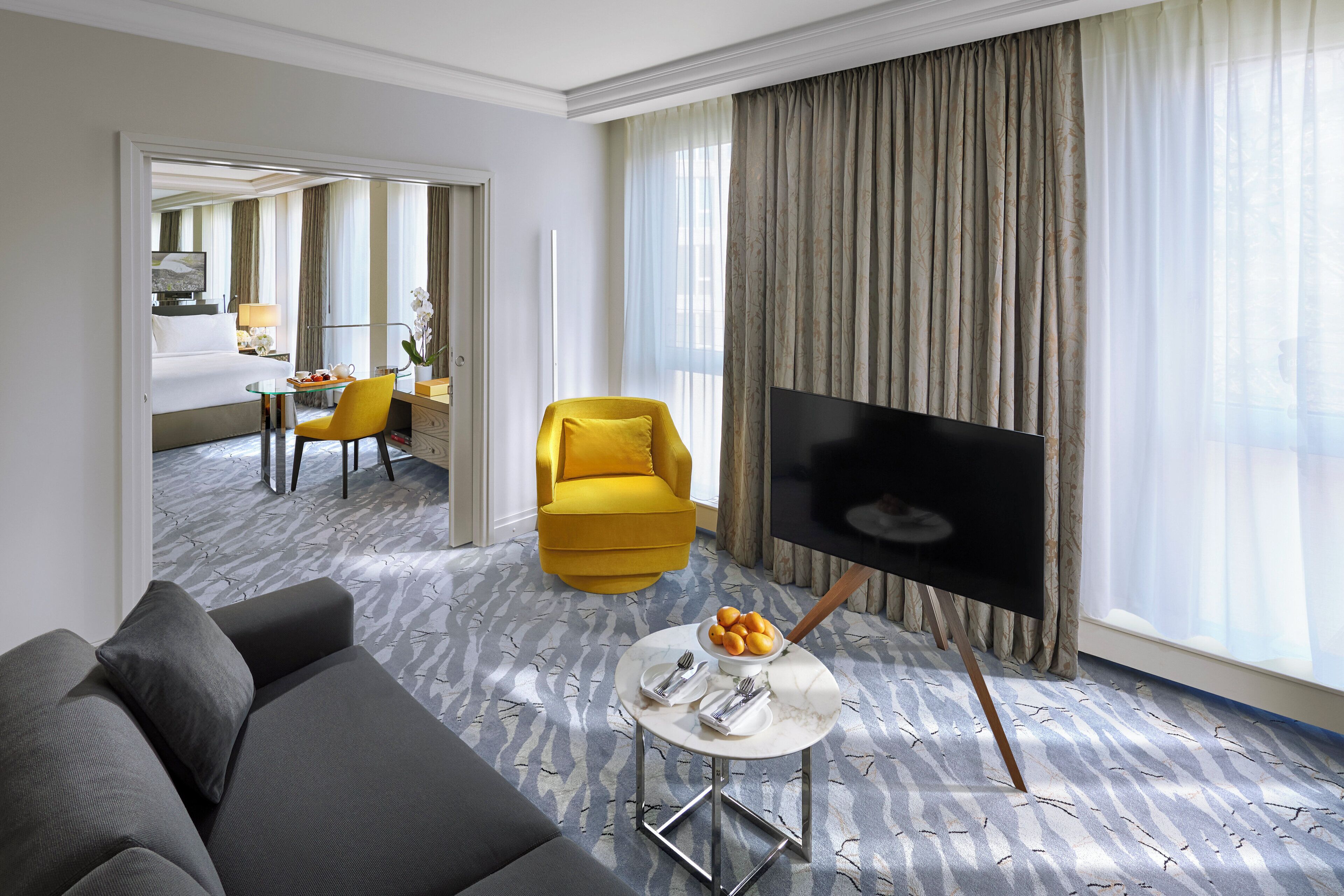 A hotel room with a sofa, a TV, a dining table with a bowl of fruit, and a chair.