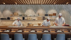 A restaurant with a marble wall, chefs preparing food, and a long dining table with chairs.